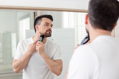 Photo of Handsome man shaving with trimmer near mirror at home