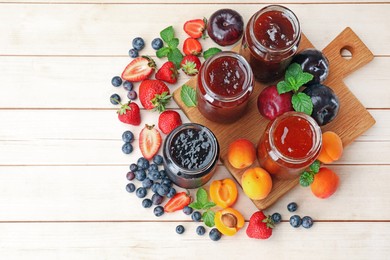 Photo of Tasty sweet jams in jars surrounded by ingredients on light wooden table, flat lay. Space for text