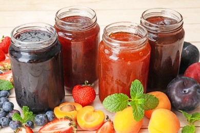 Photo of Tasty sweet jams in jars surrounded by ingredients on light wooden table, closeup