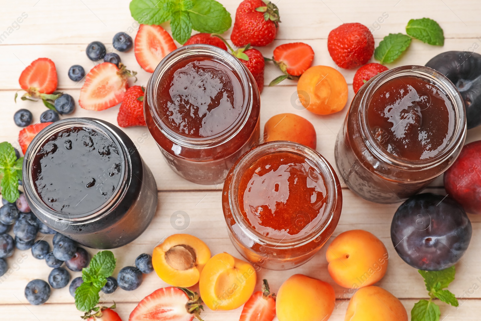 Photo of Tasty sweet jams in jars surrounded by ingredients on light wooden table, flat lay