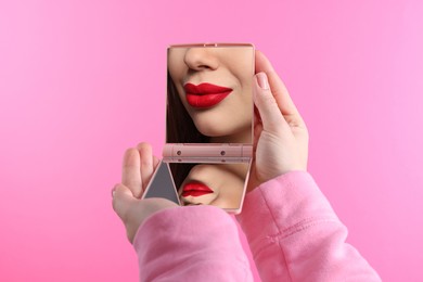 Image of Beautiful woman with red lips looking in compact mirror on pink background, closeup