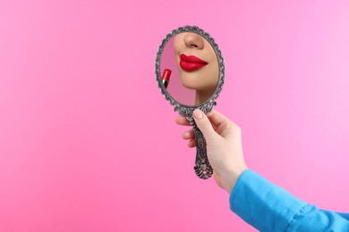 Image of Beautiful woman with red lips looking in handheld mirror on pink background, closeup