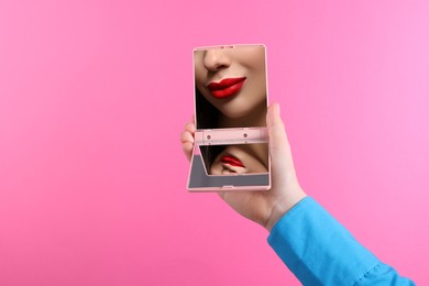 Image of Beautiful woman with red lips looking in compact mirror on pink background, closeup