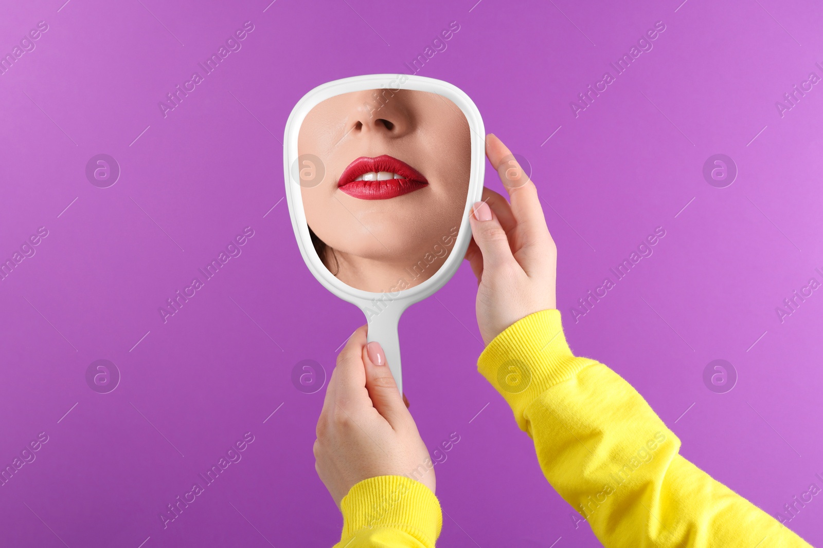 Image of Beautiful woman with red lips looking in handheld mirror on violet background, closeup