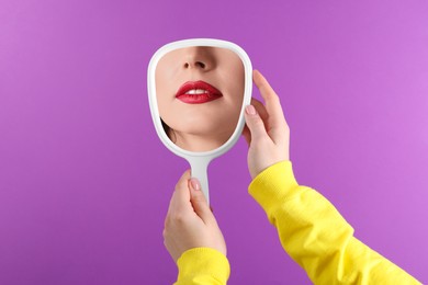 Image of Beautiful woman with red lips looking in handheld mirror on violet background, closeup