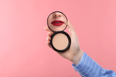 Image of Beautiful woman with red lips looking in compact powder mirror on pink background, closeup