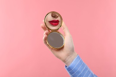 Image of Beautiful woman with red lips looking in compact mirror on pink background, closeup