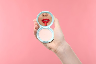 Image of Beautiful woman looking in compact powder mirror while applying red lipstick on pink background, closeup