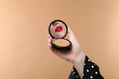 Image of Beautiful woman with red lips looking in compact powder mirror on dark beige background, closeup
