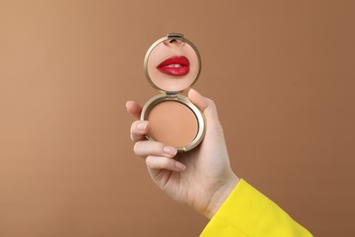 Image of Beautiful woman with red lips looking in compact powder mirror on pale brown background, closeup