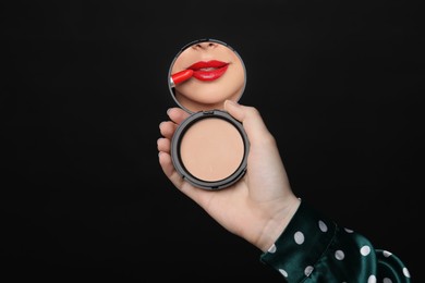 Image of Beautiful woman looking in compact powder mirror while applying red lipstick on black background, closeup