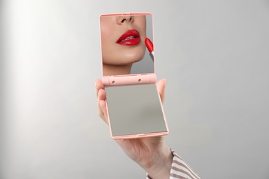 Image of Beautiful woman looking in compact mirror while applying red lipstick on light grey background, closeup