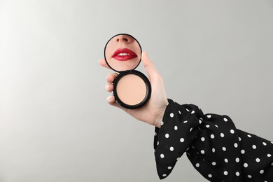 Image of Beautiful woman with red lips looking in compact powder mirror on light background, closeup