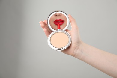 Image of Beautiful woman looking in compact powder mirror while applying red lipstick on light grey background, closeup
