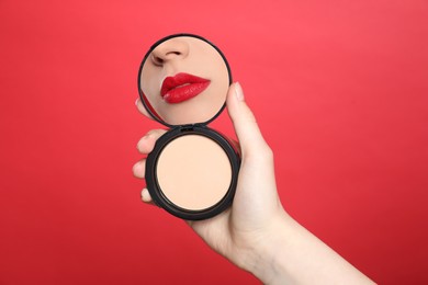 Image of Beautiful woman with red lips looking in compact powder mirror on red background, closeup