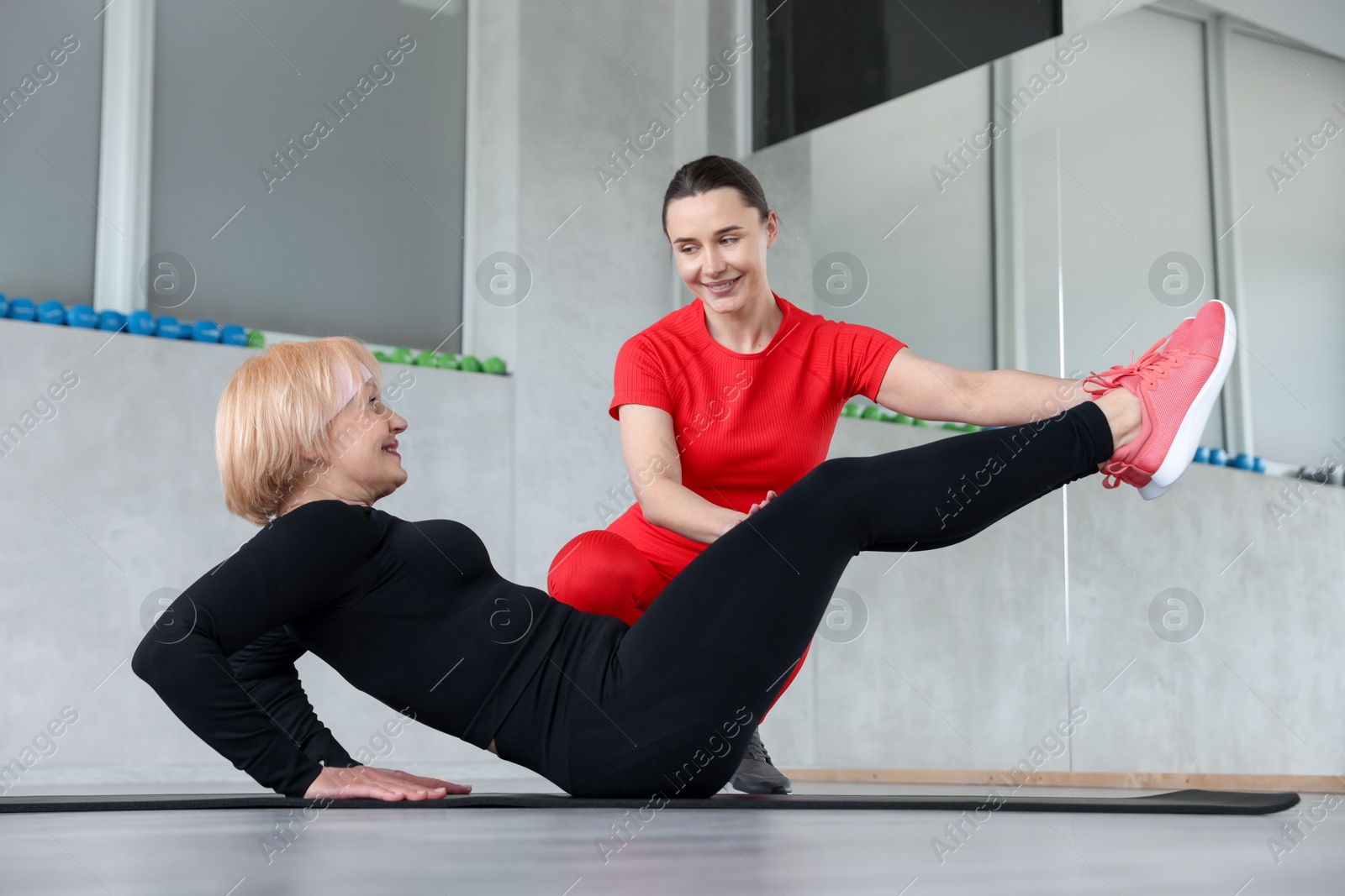 Photo of Senior woman exercising on yoga mat supervised by professional trainer in gym