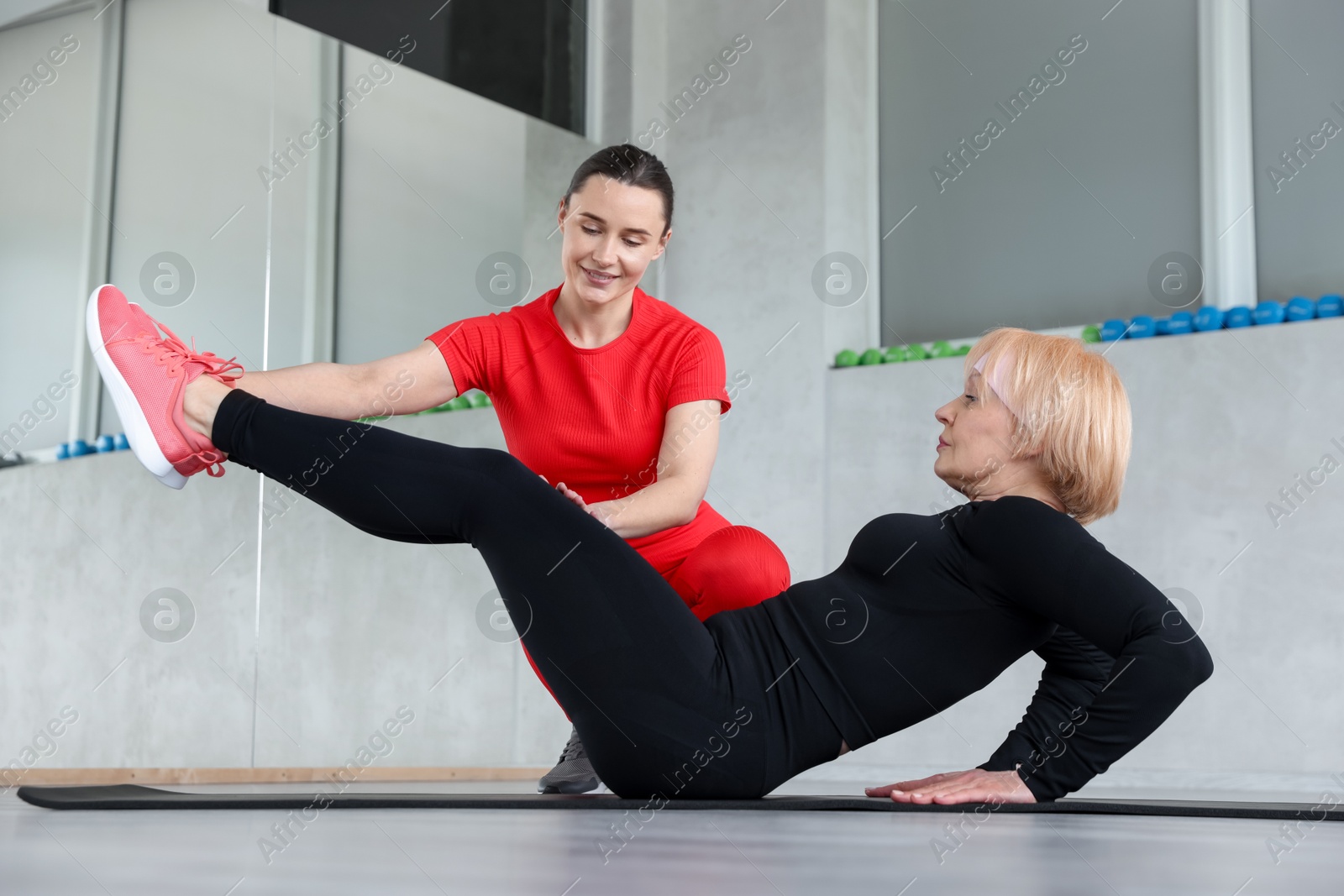Photo of Senior woman exercising on yoga mat supervised by professional trainer in gym