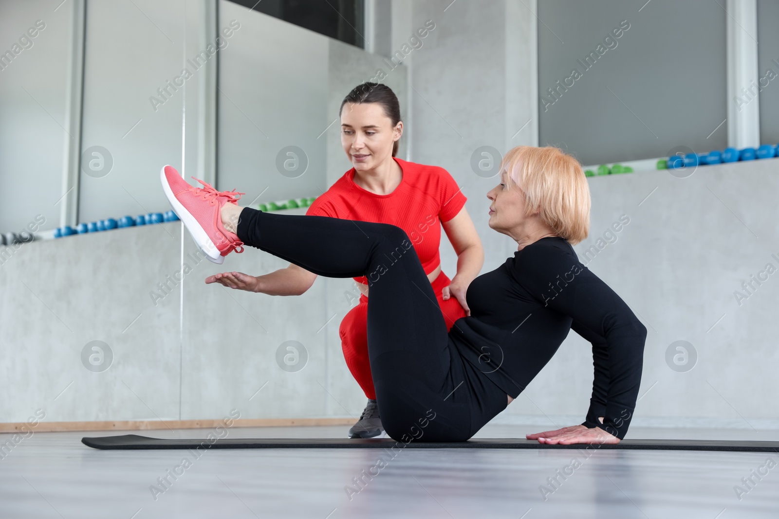 Photo of Senior woman exercising on yoga mat supervised by professional trainer in gym