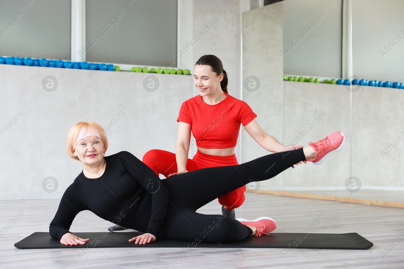 Photo of Senior woman exercising on yoga mat supervised by professional trainer in gym