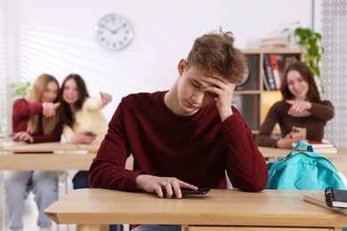Photo of Cyber bullying. Upset teenage boy with smartphone and schoolmates pointing at him in classroom, selective focus