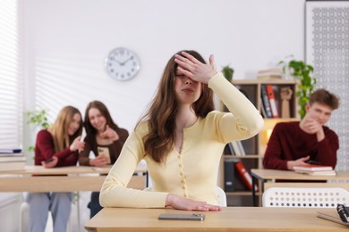 Photo of Cyber bullying. Upset teenage girl with smartphone and schoolmates pointing at her in classroom, selective focus