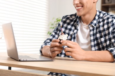 Photo of Birthday celebration. Man with cupcake having video call via laptop at table indoors, closeup