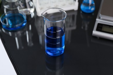 Photo of Beaker with blue liquid, other laboratory glassware and scale on black mirror table, closeup