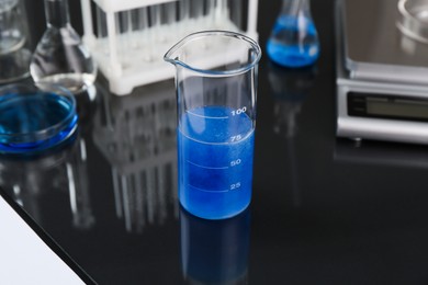 Photo of Beaker with blue liquid, other laboratory glassware and scale on black mirror table, closeup