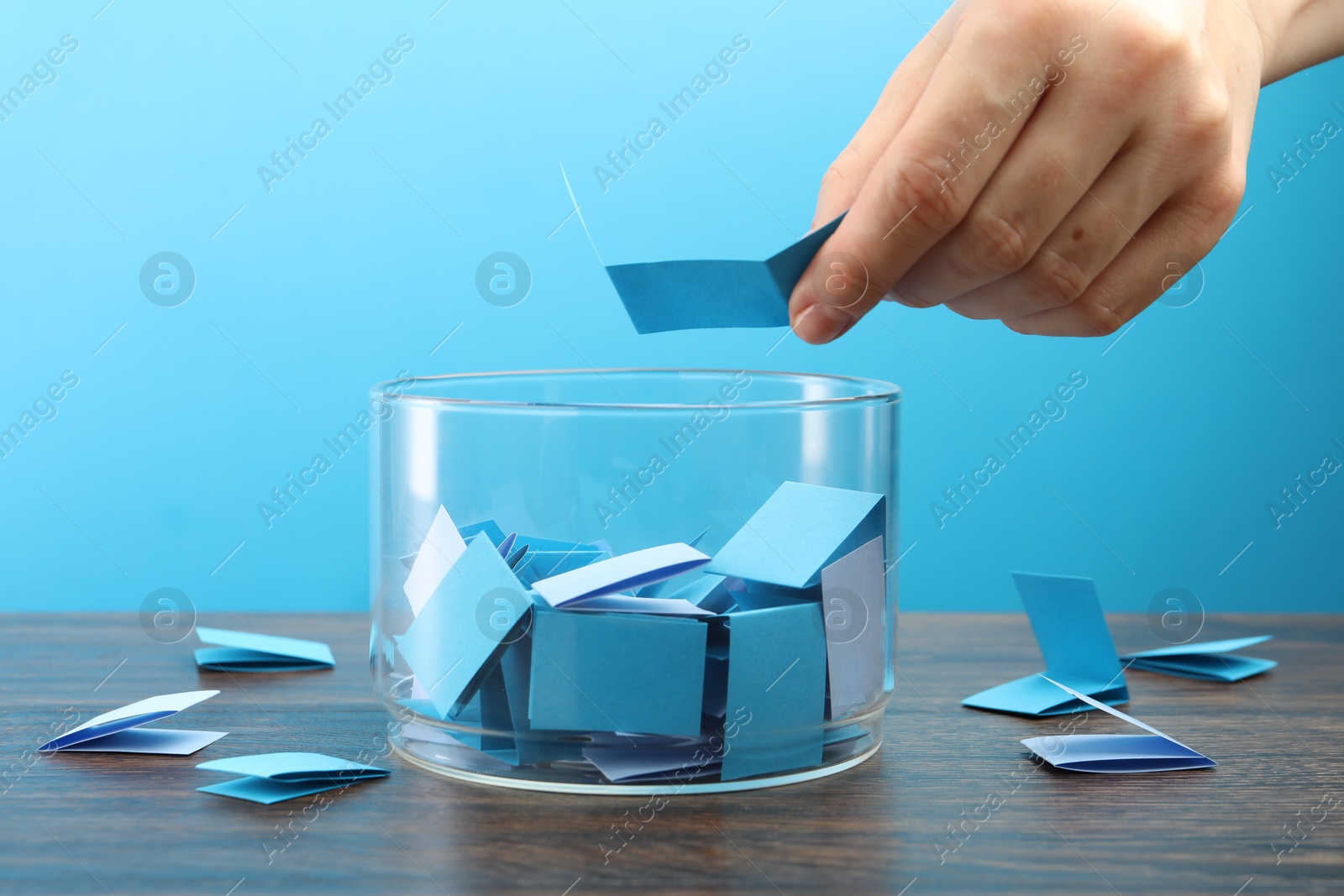 Woman taking color paper piece form glass jar at wooden table against light blue background, closeup Photo of Woman taking color paper piece form glass jar at wooden table against light blue background, closeup