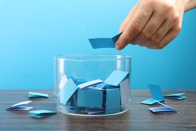 Photo of Woman taking color paper piece form glass jar at wooden table against light blue background, closeup