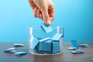 Photo of Woman taking color paper piece form glass jar at wooden table against light blue background, closeup