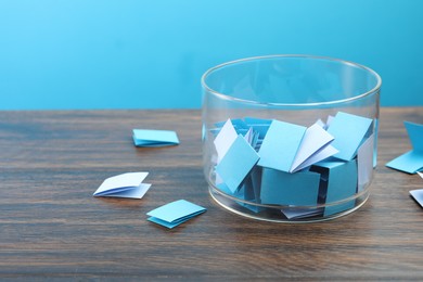 Photo of Colorful paper pieces in glass jar on wooden table against light blue background, closeup. Space for text