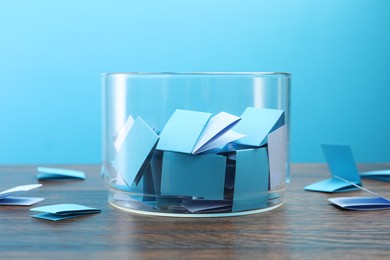 Photo of Colorful paper pieces in glass jar on wooden table against light blue background, closeup