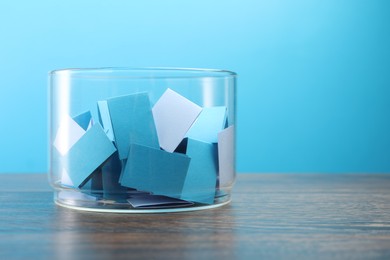 Photo of Colorful paper pieces in glass jar on wooden table against light blue background, space for text