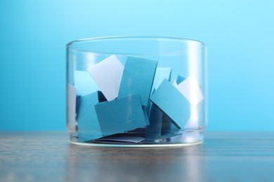 Photo of Colorful paper pieces in glass jar on wooden table against light blue background, closeup