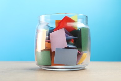 Photo of Colorful paper pieces in glass jar on wooden table against light blue background