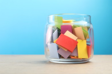 Photo of Colorful paper pieces in glass jar on wooden table against light blue background, space for text