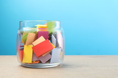 Photo of Colorful paper pieces in glass jar on wooden table against light blue background, space for text