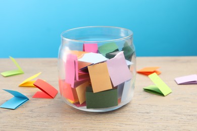 Photo of Colorful paper pieces in glass jar on wooden table against light blue background, closeup