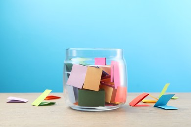 Photo of Colorful paper pieces in glass jar on wooden table against light blue background