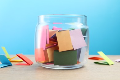 Photo of Colorful paper pieces in glass jar on wooden table against light blue background, closeup