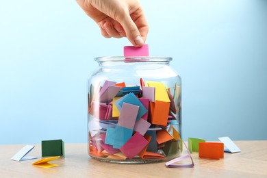 Photo of Woman taking color paper piece form glass jar at wooden table against light blue background, closeup
