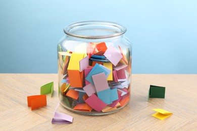 Photo of Colorful paper pieces in glass jar on wooden table against light blue background