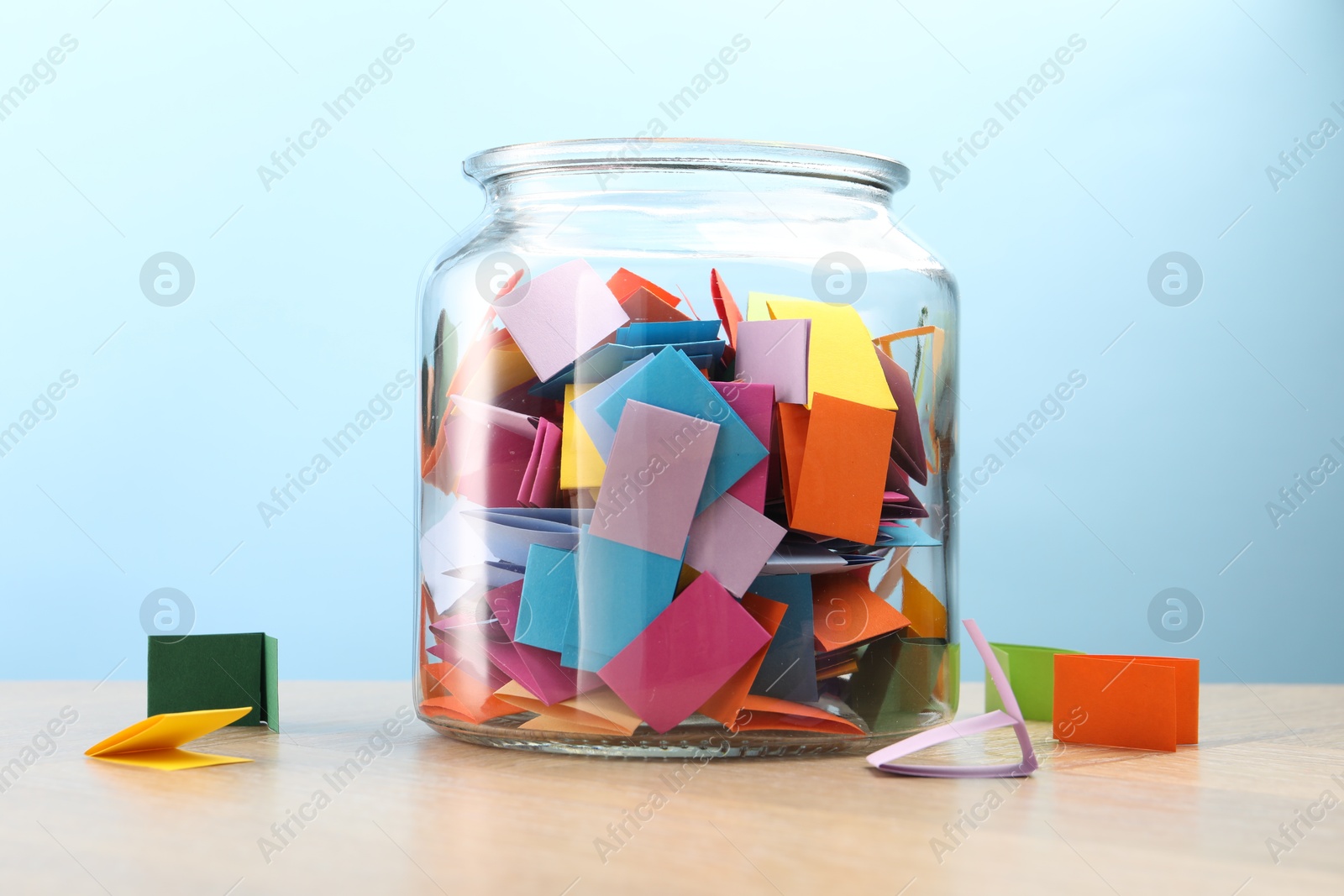 Colorful paper pieces in glass jar on wooden table against light blue background Photo of Colorful paper pieces in glass jar on wooden table against light blue background