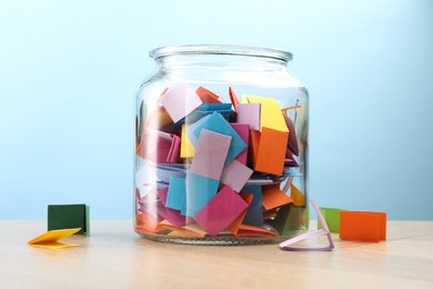 Photo of Colorful paper pieces in glass jar on wooden table against light blue background