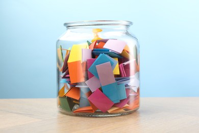 Photo of Colorful paper pieces in glass jar on wooden table against light blue background