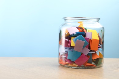 Photo of Colorful paper pieces in glass jar on wooden table against light blue background, space for text