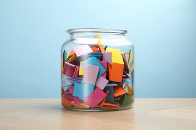 Photo of Colorful paper pieces in glass jar on wooden table against light blue background