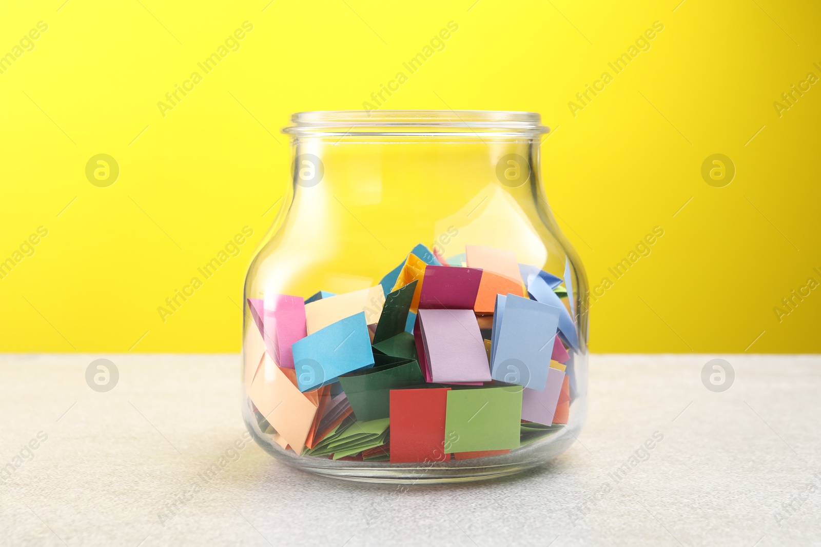 Jar with colorful pieces of paper on light table against yellow background Photo of Jar with colorful pieces of paper on light table against yellow background