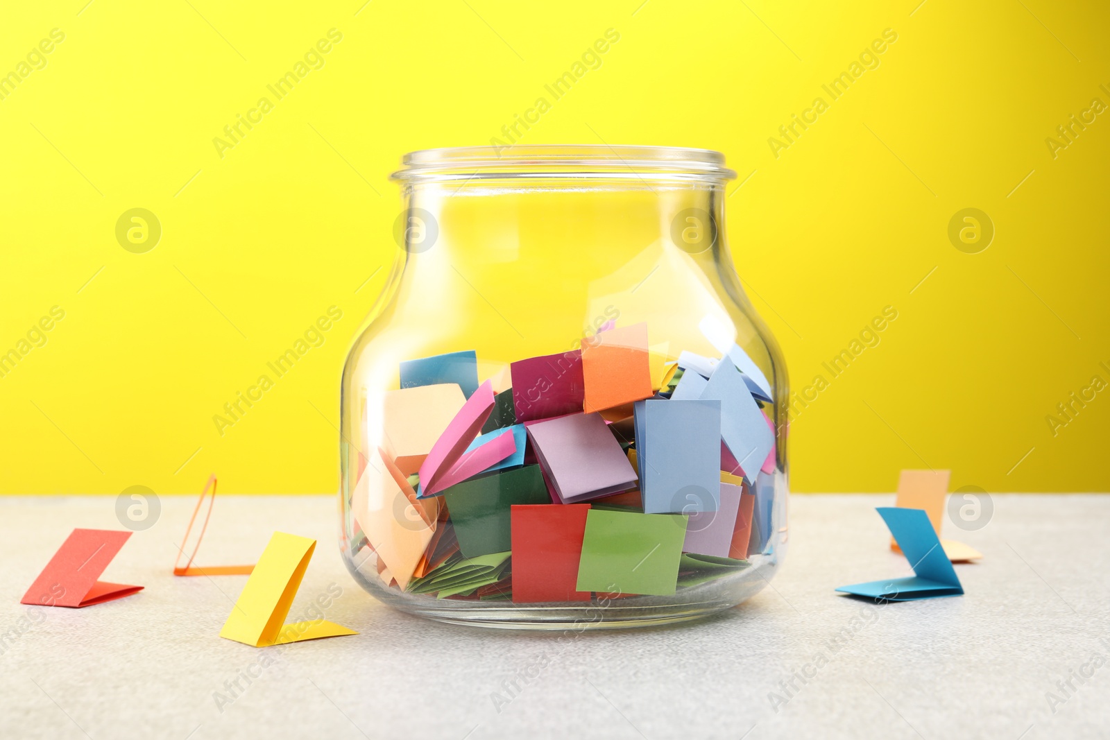 Jar with colorful pieces of paper on light table against yellow background Photo of Jar with colorful pieces of paper on light table against yellow background